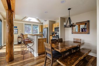 A dining area with a long wooden table and chairs, a breakfast bar with high stools, and a kitchen in the background.