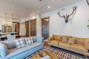 The living room of Modern Blackstone Paradise 2A, with a light gray sofa and a tan leather sofa, an antler wall hanging, and a view into the kitchen and dining area.