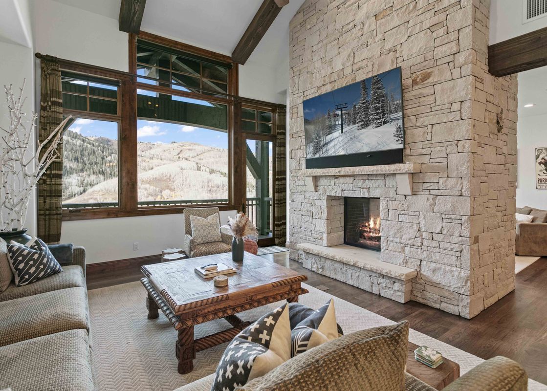 A spacious living area featuring a stone fireplace with a mounted television above it, surrounded by comfortable sofas and a rustic coffee table, all looking out to snow-covered mountain views.