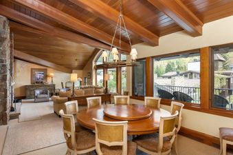 The dining area in Plaza Lodge 5 Bedroom Gold Unit 7 features a large round wooden table with seating for eight, illuminated by a rustic chandelier, and offers expansive views of the outdoors through large windows.