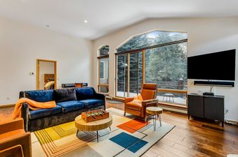 A modern living room with large windows overlooking snow-covered trees, featuring a blue velvet sofa, a leather armchair, a geometric rug, and a foosball table.