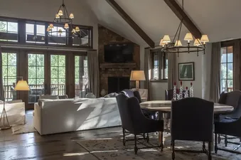 An expansive open-concept living and dining area within Martin Creek Cottage, featuring a stone fireplace, large windows, a white sofa, and a round dining table with upholstered chairs.