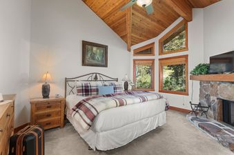 The bedroom in Crescent Cabin features a large bed with plaid bedding, a stone fireplace, and multiple windows offering views of the surrounding nature.