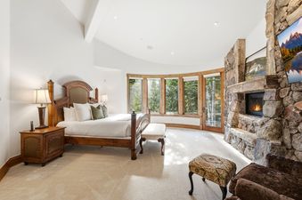 Spacious master bedroom at Northside Retreat with a four-poster wooden bed, large stone fireplace, and curved bay windows overlooking the trees.