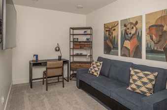 A modern and cozy den area features a blue sofa, a dark wooden desk with a leather chair, a tall metal bookshelf, and three abstract animal portraits on the wall.