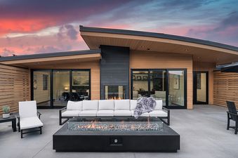 An expansive outdoor patio with a large fire pit, comfortable seating, and a dining area, set against the backdrop of a modern lodge at sunset.