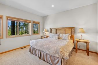 A cozy bedroom at Wood Run Ski Haus features a large bed with a wooden headboard, two nightstands with lamps, and windows overlooking a vibrant evergreen forest.