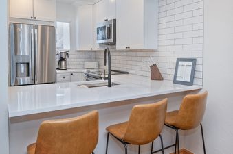 A kitchen with white cabinets, a stainless steel refrigerator, and a breakfast bar with brown leather stools.