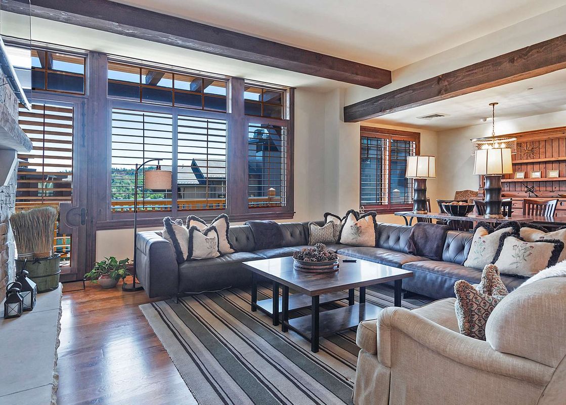 A luxurious living room features a large sectional sofa, a coffee table with pinecones, a stone fireplace, and large windows with wooden blinds revealing an exterior view.