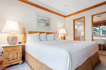 A cozy bedroom with a king-sized bed, two nightstands with lamps, a framed painting, and an open doorway leading to a bathroom.
