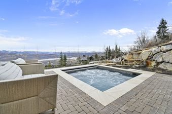 The hot tub at the Deer Crest Escape Retreat features bubbling water and is surrounded by a paved patio, offering stunning views of mountains and valleys.
