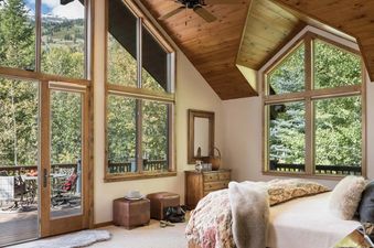 A cozy bedroom with a wooden vaulted ceiling, a comfortable bed, and large windows looking out onto a private balcony and a forest.