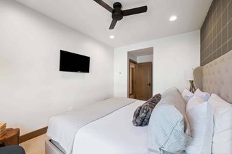 The bedroom at Village Hall 13 features a comfortable bed, a wall-mounted television, and a modern ceiling fan, ensuring a relaxing stay.