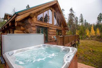 A rustic log cabin with a large wooden deck and a bubbling hot tub in the foreground, surrounded by an autumn forest.