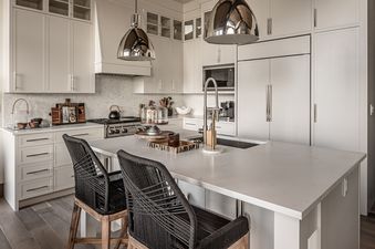 A modern kitchen in Inspiration Point Townhome 4C features white cabinetry, a large white quartz island with two bar stools, stainless steel appliances, and polished silver pendant lights.