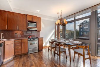 A modern kitchen and dining area in Lowell 402A features wooden cabinetry, red granite countertops, stainless steel appliances, and a glass-top dining table with chairs, all illuminated by natural light from large windows offering mountain views.