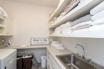 A clean and organized laundry room at Turkey Creek with a washing machine, sink, and shelves stocked with neatly folded towels.