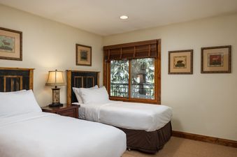 A cozy bedroom at Montaneros 418 with two twin beds, dark wooden headboards, a bedside lamp, framed animal artworks, and a window offering a view of green trees.