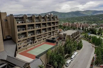 An aerial view showcases the One Bedroom Suite Beaver Run Resort's Brutalist architecture, with its tiered structure and many windows, a tennis court at its base, and surrounding green mountains under a cloudy sky.