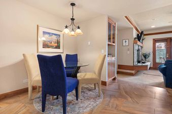 The dining area in The Charter C0405 features a glass dining table with blue and beige chairs, illuminated by a chandelier and decorated with a wall painting.