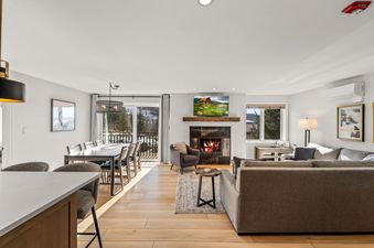 A bright open-concept living and dining area with light wood floors, a cozy fireplace, a large sofa, a dining table, and a balcony with snowy outdoor views.