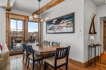 A rustic-chic dining room with a large wooden table, surrounded by six chairs, under a contemporary chandelier, with a large artwork of a snowy mountain scene on the wall, and French doors leading to a balcony.