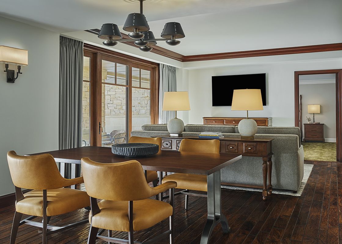 A dining table with four chairs and a decorative bowl sits in the foreground, with a living area featuring a sofa, lamps, and a television visible in the background, all within the Montage Deer Valley Accessible Alpine Residential Suite with Fire Pit.