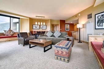 The living area of The Grand Summit Lodge Studio Residence features a patterned rug, a gray couch with throw pillows, a dark brown armchair, and a wooden coffee table, with a dining area and kitchen in the background.