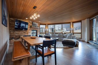 A wide shot of Chateau Chamonix CX111's living and dining areas, featuring a wooden dining table, a sectional sofa, a fireplace, and large windows with an outdoor view.