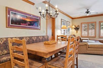 The dining area in The Charter D2505 features a rustic wooden table, an upholstered bench, and wooden chairs, all set under a chandelier and next to a large framed landscape artwork.