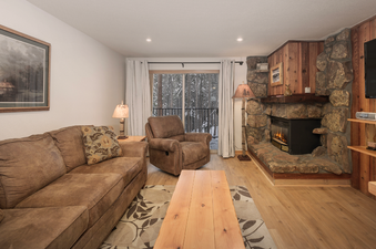 A cozy living room at Beaver Village 1612 features a brown sofa, a matching recliner, a rustic stone fireplace with an electric insert, and a large window leading to a snowy outdoor view.