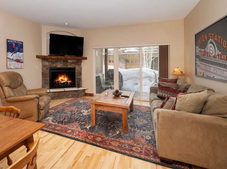 A cozy living room in Telemark 514 features a stone fireplace, a large TV, and comfortable seating, with a view of a snowy outdoor patio.