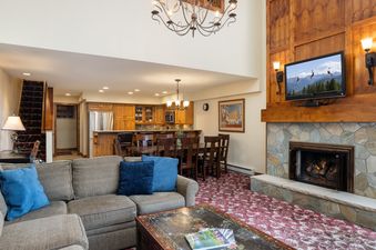 A cozy living room at Montaneros 418 featuring a sectional sofa, a stone fireplace with a TV above it, and a dining area connected to the kitchen.
