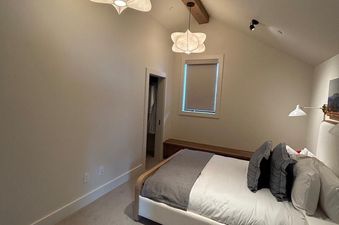 The bedroom in Moonlight Basin 7 Deep Forest has a large, comfortable bed with a gray throw blanket, white linens, and white and gray pillows. The bed is against a wall with a bedside table and a window to the left. The ceiling has exposed wooden beams a