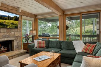 A cozy living room with a green sectional sofa, a stone fireplace, and large windows looking out onto a lush forest.