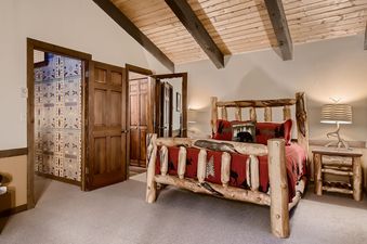 The bedroom at Breck Lodge features a rustic log bed with red bedding and accent pillows, set against a light-colored wall with a vaulted wood-paneled ceiling.