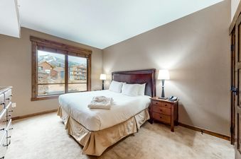 The master bedroom at Trailhead Lodge 2105 with a king-sized bed, nightstands, lamps, and a large window offering mountain and lodge views.