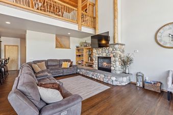 A spacious living room at Wood Run Ski Haus with an L-shaped sectional sofa, a stone fireplace, a wall-mounted TV, and a rustic wooden bookshelf.