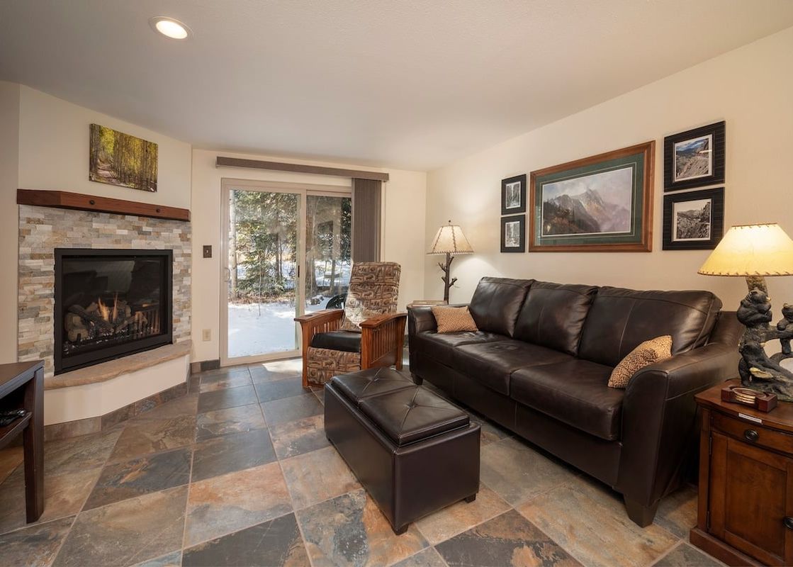 A cozy living room at Beaver Village 414 features a brown leather sofa, stone fireplace, and access to a snowy outdoor area.