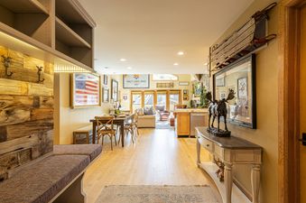 A wide interior shot of the Royal Plaza 403, showing the dining area with a long wooden table and chairs, leading into a living space with comfortable seating, decorative art, and a view towards large windows, with hardwood floors throughout.