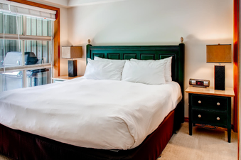 A cozy bedroom featuring a comfortable bed with white linens, a dark green headboard, and wooden accents, bathed in natural light from a large window.