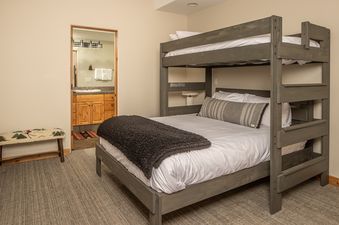 A bunk room at Moonlight Mountain 8 Happy Trails features a gray wooden bunk bed with white bedding and a dark gray throw, a decorative bench, and an en suite bathroom with a wooden vanity and mirror.