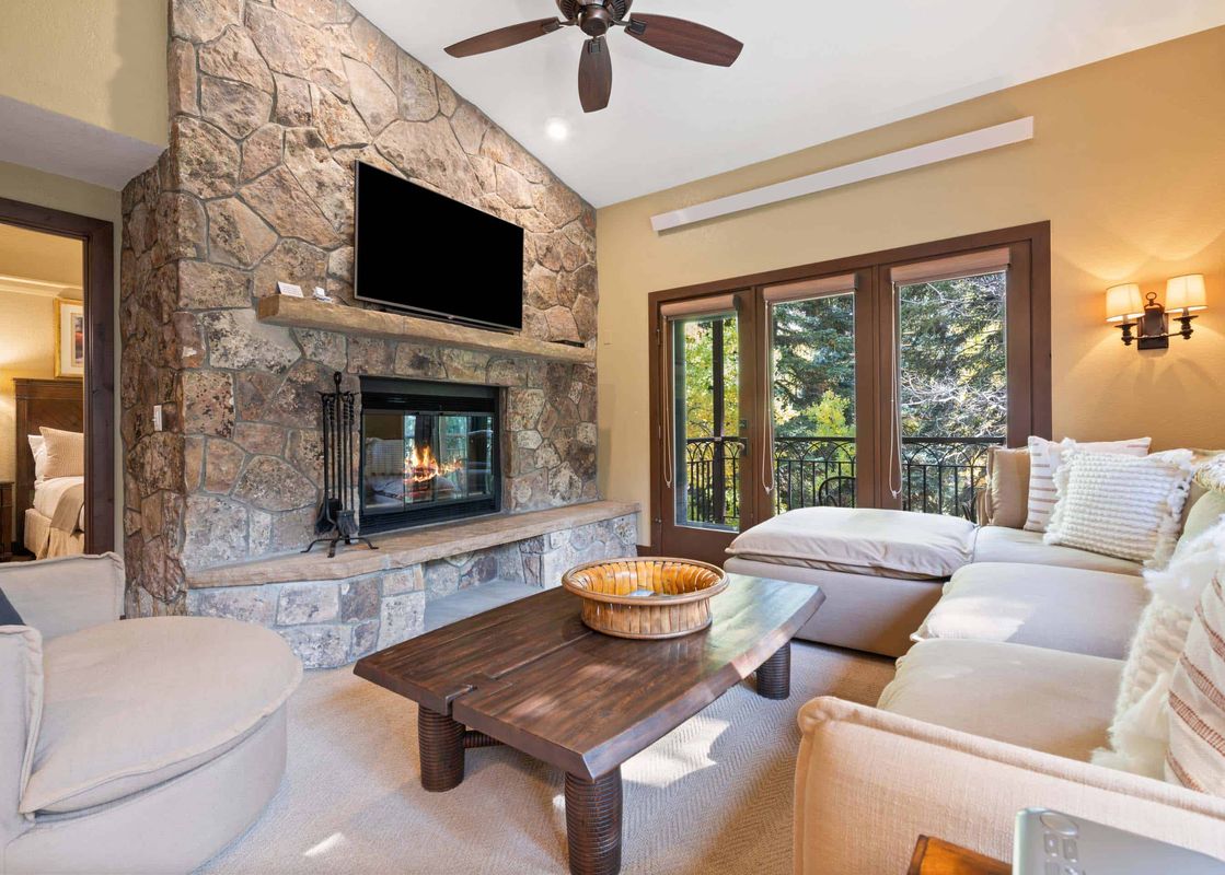 The living area of The Charter C2105 features a large stone fireplace, a wall-mounted television, comfortable sofas, a wooden coffee table, and windows overlooking a wooded landscape.
