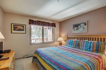 A bedroom with a wooden bed, a striped blue and yellow blanket, and two windows with a view of the trees outside