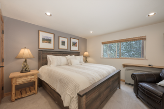 A master bedroom in Beaver Village 1612 features a large bed with a dark wood frame and white bedding, a wooden nightstand with a lamp, and a window offering a snowy outdoor view.
