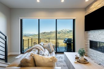 A luxurious living room with a large leather sofa, a modern coffee table, a fireplace, and a stunning view of mountains and a lake through glass sliding doors at Hideout Epic View.