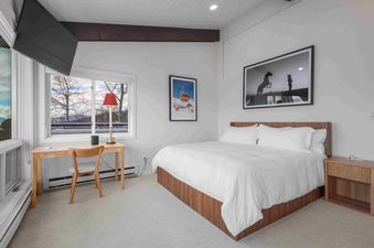 The image shows the bedroom in Enclave 313, featuring a bed with white linens, a wooden desk by a window with a snowy mountain view, and two framed art pieces on the wall.