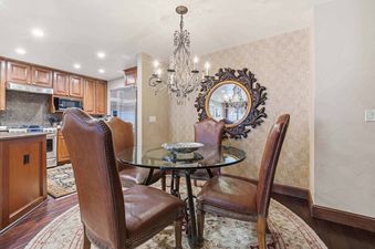 A dining area with a round glass-top table, brown leather chairs, and a chandelier, adjacent to a kitchen with wooden cabinets and stainless steel appliances.