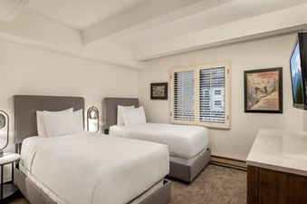 A cozy bedroom with two twin beds, a bedside table with a lamp, and a window with shutters.