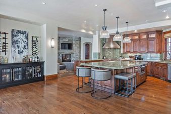 A spacious kitchen at Alpenglow at the Colony featuring a large island with bar stools, rich wooden cabinetry, green granite countertops, and an adjacent bar area, with a stone fireplace visible in the background.